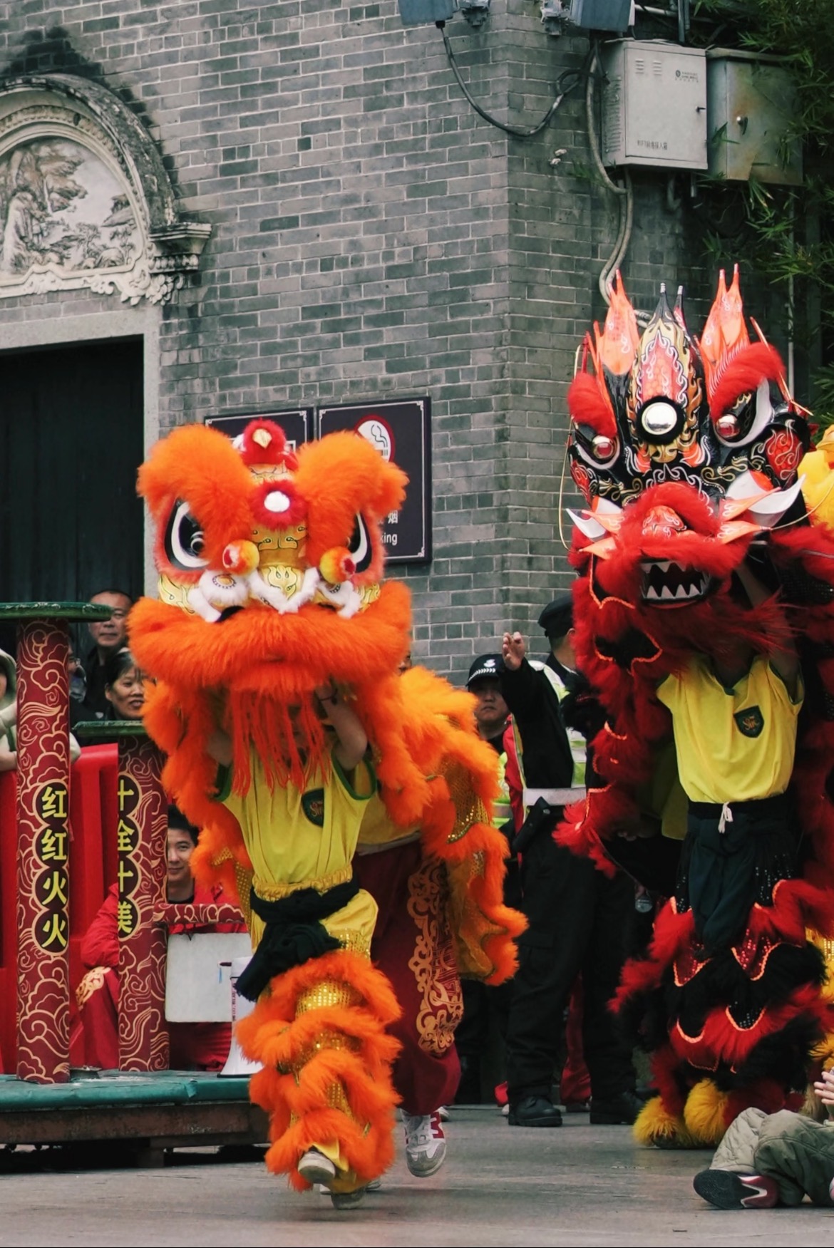 橙色与黑红色醒狮同台表演,背景为岭南青砖街道,气氛热烈